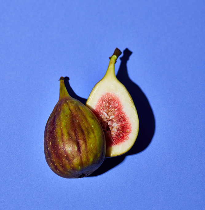 Eine halbierte Feige wird vor einem violetten Hintergrund präsentiert, fachmännisch arrangiert von einem Foodstylisten. Die eine Hälfte der Feige hat eine grün-violett gestreifte Schale, während die andere Hälfte ihr rosa, mit Samen gefülltes Inneres offenbart. Die kontrastierenden Farben lassen die Früchte in dieser atemberaubenden Lebensmittelfotografie hervorstechen.