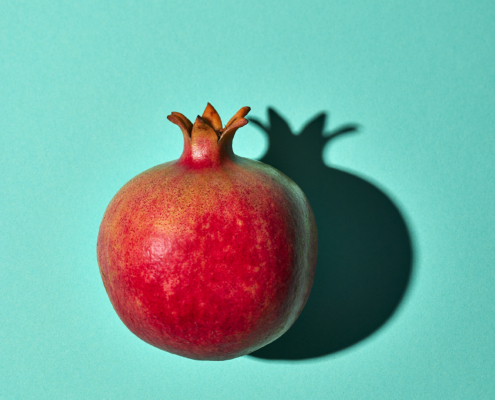 Ein ganzer Granatapfel mit rot und rosa gesprenkelter Schale liegt auf einem hellen türkisfarbenen Hintergrund. Der obere Kronenabschnitt des Granatapfels ist deutlich sichtbar und wirft einen Schatten nach rechts. Diese minimalistische und lebendige Komposition zeigt exquisite Fotografie Lebensmittel und hebt die Textur und Farbe der Frucht hervor.