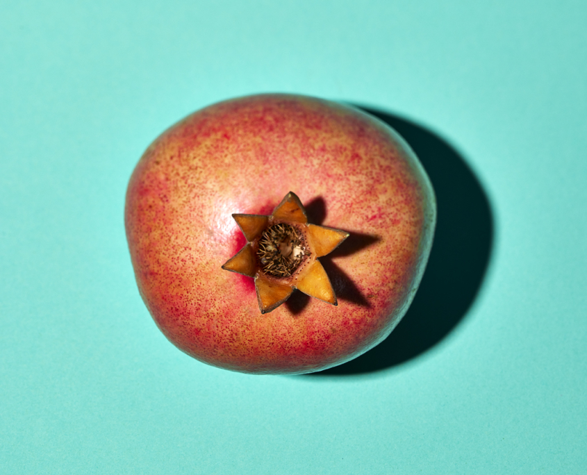 Ein reifer Granatapfel mit leuchtend roter Außenschale liegt auf einer mintgrünen Oberfläche, wunderschön eingefangen von einem professionellen Foodstylisten. Die Frucht wirft einen deutlichen Schatten nach rechts, was ihre runde Form betont. Die schlichte Farbe des Hintergrunds kontrastiert mit dem Granatapfel und lässt ihn in der Bildsprache hervorstechen.