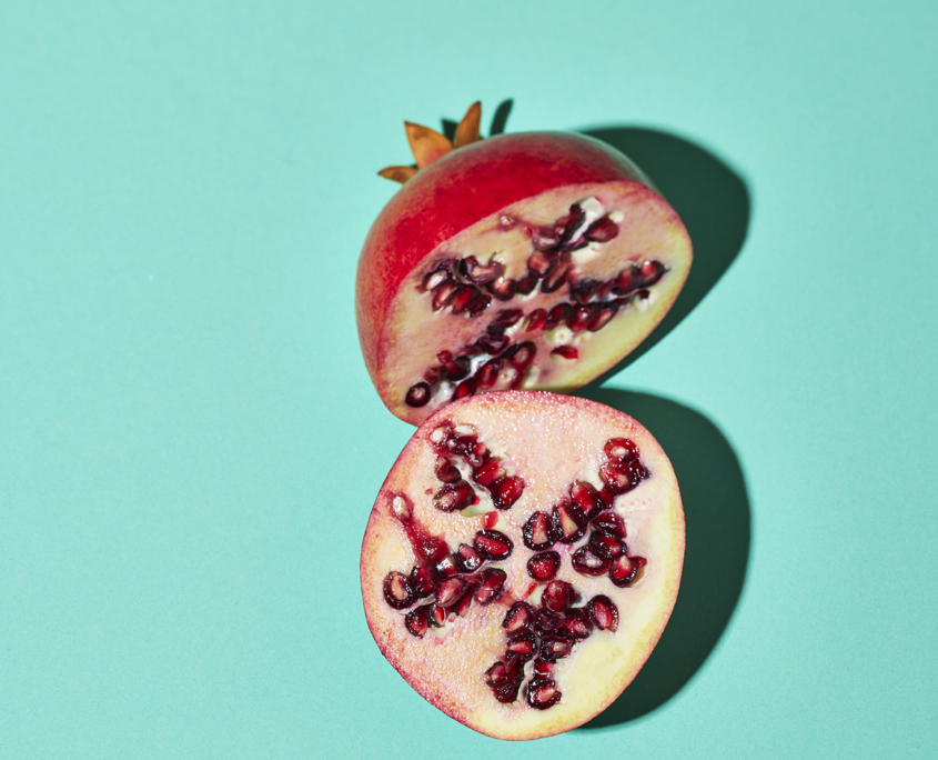 Ein halbierter Granatapfel auf einem mintgrünem Hintergrund. Die obere Hälfte der Frucht zeigt ihr Äußeres, während die untere Hälfte die saftigen roten Kerne im Inneren zeigt. Der Schatten der Frucht wird auf die mintgrüne Oberfläche geworfen und schafft so ein exquisites Beispiel für die Kunst der Foodfotografie.