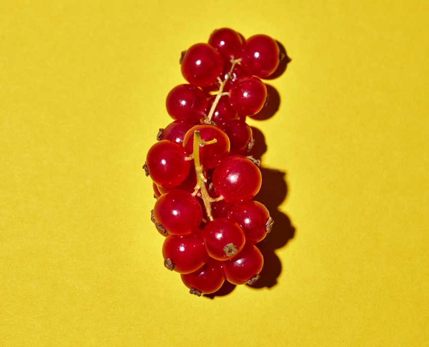 Eine kleine Traube leuchtend roter Johannisbeeren steht vor einem leuchtend gelben Hintergrund. Die Beeren stehen dicht beieinander und bilden einen optisch auffälligen Kontrast zum lebhaften Hintergrund – ein perfektes Beispiel für Food-Fotografie vom Feinsten.