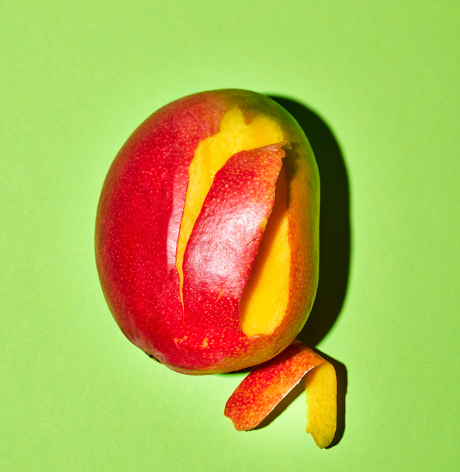 Der leuchtend grüne Hintergrund lässt das leuchtend gelbe Fruchtfleisch der teilweise geschälten Mango hervorstechen, wobei sich die rote und gelbe Schale nach außen wölbt und mehr von dem saftigen Inneren freigibt – ein perfektes Beispiel für Foodfotografie, die die Essenz der Bildsprache einfängt.