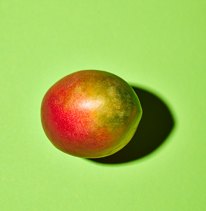 Eine reife Mango mit roter und grüner Schale liegt auf einem hellgrünen Hintergrund. Das Licht wirft einen leichten Schatten und verleiht der lebendigen Komposition, die mit der Präzision eines erfahrenen Food-Fotografen gestaltet wurde, Tiefe.