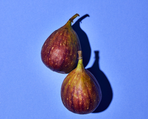 Zwei reife Feigen mit violett-grüner Schale und glatter Textur liegen auf einem leuchtend blauen Hintergrund. Die Beleuchtung erzeugt deutliche Schatten, die ihre Form und satte Farbe betonen und die komplexe Bildsprache verkörpern, die Food-Fotografen in der Foodfotografie anstreben.