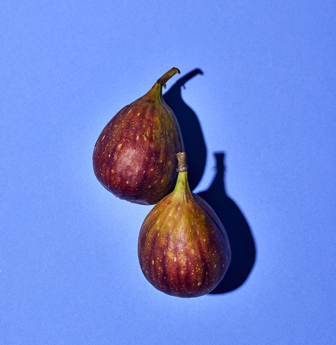Zwei reife Feigen mit violett-grüner Schale und glatter Textur liegen auf einem leuchtend blauen Hintergrund. Die Beleuchtung erzeugt deutliche Schatten, die ihre Form und satte Farbe betonen und die komplexe Bildsprache verkörpern, die Food-Fotografen in der Foodfotografie anstreben.