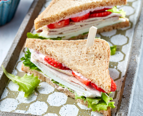Zwei dreieckige Putensandwiches mit Salat und geschnittenen Erdbeeren, mit Holzspießen befestigt, auf einem grauen rechteckigen Tablett mit gemustertem Boden. Im Hintergrund ist teilweise eine kleine Schale mit ganzen Erdbeeren zu sehen. Sie zeigt exquisite Food-Fotografie, die die Kunstfertigkeit des Food-Stylisten hervorhebt.