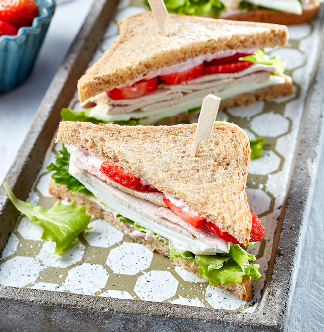 Zwei dreieckige Putensandwiches mit Salat und geschnittenen Erdbeeren, mit Holzspießen befestigt, auf einem grauen rechteckigen Tablett mit gemustertem Boden. Im Hintergrund ist teilweise eine kleine Schale mit ganzen Erdbeeren zu sehen. Sie zeigt exquisite Food-Fotografie, die die Kunstfertigkeit des Food-Stylisten hervorhebt.