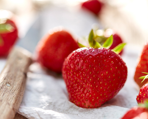 Nahaufnahme von reifen, roten Erdbeeren mit grünen Blättern auf einer weißen strukturierten Oberfläche. Die Erdbeeren sind im Fokus, während der Hintergrund leicht verschwommen ist, was einen Bokeh-Effekt erzeugt. Auf der linken Seite des Bildes ist leicht ein Holzobjekt zu erkennen, das exquisite Food-Fotografie zeigt.