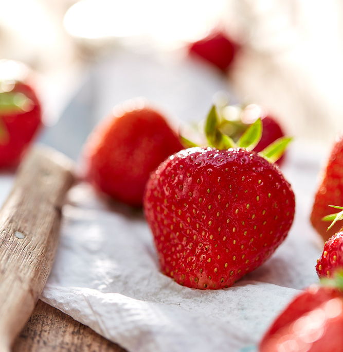 Nahaufnahme von reifen, roten Erdbeeren mit grünen Blättern auf einer weißen strukturierten Oberfläche. Die Erdbeeren sind im Fokus, während der Hintergrund leicht verschwommen ist, was einen Bokeh-Effekt erzeugt. Auf der linken Seite des Bildes ist leicht ein Holzobjekt zu erkennen, das exquisite Food-Fotografie zeigt.