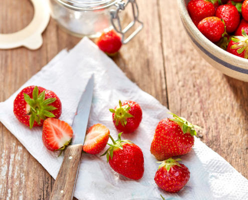 Auf einem Holztisch stehen ein Glas, eine Keramikschale mit Erdbeeren und ein paar Erdbeeren auf einem weißen Papiertuch. Auf dem Tuch liegt ein Messer mit Holzgriff, in dem eine Erdbeere halbiert ist, sodass man ihr Inneres sehen kann. Die Bildsprache fängt die Essenz der Foodfotografie wunderbar ein.
