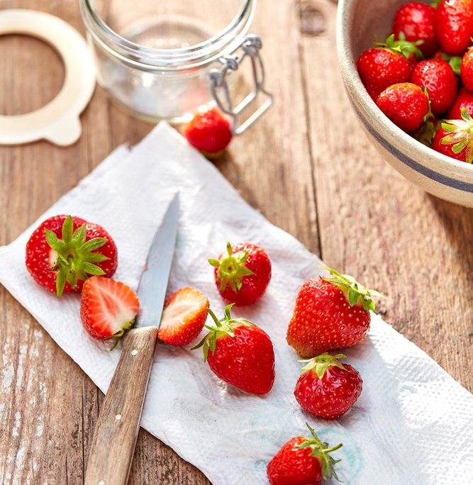 Auf einem Holztisch stehen ein Glas, eine Keramikschale mit Erdbeeren und ein paar Erdbeeren auf einem weißen Papiertuch. Auf dem Tuch liegt ein Messer mit Holzgriff, in dem eine Erdbeere halbiert ist, sodass man ihr Inneres sehen kann. Die Bildsprache fängt die Essenz der Foodfotografie wunderbar ein.