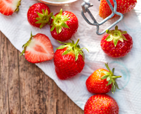 Auf einer Holzoberfläche werden frische Erdbeeren und ein Glas mit braunem Zucker präsentiert. Die Erdbeeren sind ganz und halbiert und liegen auf einem weißen Papiertuch. Der Deckel des Glases ist teilweise geöffnet, im Inneren sind einige Zuckerkörner zu sehen. Natürliches Sonnenlicht erhellt die Szene und veranschaulicht exquisite Fotografie Lebensmittel.
