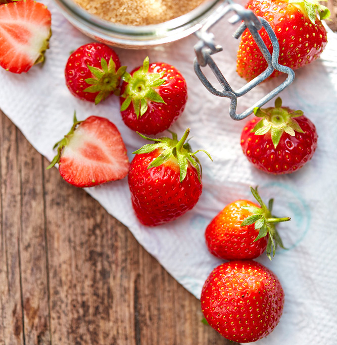 Auf einer Holzoberfläche werden frische Erdbeeren und ein Glas mit braunem Zucker präsentiert. Die Erdbeeren sind ganz und halbiert und liegen auf einem weißen Papiertuch. Der Deckel des Glases ist teilweise geöffnet, im Inneren sind einige Zuckerkörner zu sehen. Natürliches Sonnenlicht erhellt die Szene und veranschaulicht exquisite Fotografie Lebensmittel.