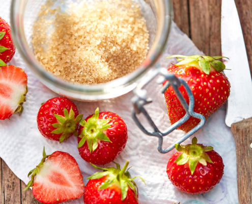 Ein Glas brauner Zucker neben mehreren frischen Erdbeeren, einige davon halbiert, ergibt ein perfektes Foodfotografie-Tableau. Die Szene ist auf einer Holzoberfläche mit einem weißen Tuch unter den Erdbeeren und einem Messer auf der rechten Seite aufgebaut und fängt die Essenz des sorgfältigen Food-Stylings ein.