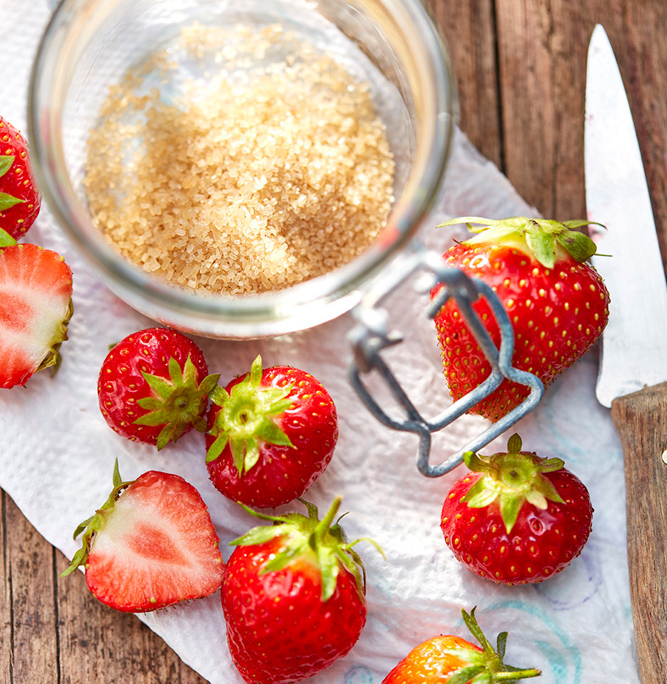 Ein Glas brauner Zucker neben mehreren frischen Erdbeeren, einige davon halbiert, ergibt ein perfektes Foodfotografie-Tableau. Die Szene ist auf einer Holzoberfläche mit einem weißen Tuch unter den Erdbeeren und einem Messer auf der rechten Seite aufgebaut und fängt die Essenz des sorgfältigen Food-Stylings ein.