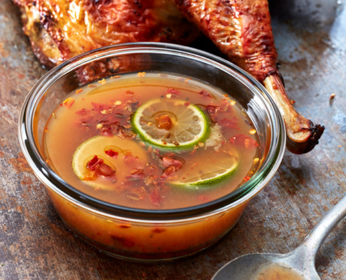 Ein gebratenes, goldbraunes Hähnchen ruht auf einer rustikalen Metalloberfläche mit einer kleinen Glasschale mit Chili-Limetten-Dip-Sauce im Vordergrund. Die Sauce enthält Limettenscheiben und Chiliflocken. Ein Löffel mit Saucenresten liegt daneben und fängt die Essenz der Food-Fotografie perfekt ein.