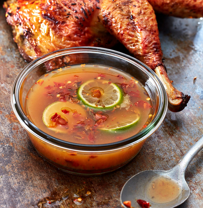Ein gebratenes, goldbraunes Hähnchen ruht auf einer rustikalen Metalloberfläche mit einer kleinen Glasschale mit Chili-Limetten-Dip-Sauce im Vordergrund. Die Sauce enthält Limettenscheiben und Chiliflocken. Ein Löffel mit Saucenresten liegt daneben und fängt die Essenz der Food-Fotografie perfekt ein.
