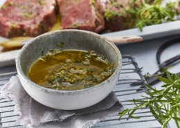Eine kleine Schüssel mit einer Marinade aus grünen Kräutern steht auf einem Tuch auf einem Metallgitter zum Abkühlen. Im Hintergrund liegt ein mariniertes rohes Steak auf einer weißen Platte, perfekt eingefangen durch professionelle Food-Fotografie. Frische Kräuter sind überall verstreut und unterstreichen das rustikale kulinarische Ambiente.