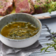 Eine kleine Schüssel mit einer Marinade aus grünen Kräutern steht auf einem Tuch auf einem Metallgitter zum Abkühlen. Im Hintergrund liegt ein mariniertes rohes Steak auf einer weißen Platte, perfekt eingefangen durch professionelle Food-Fotografie. Frische Kräuter sind überall verstreut und unterstreichen das rustikale kulinarische Ambiente.