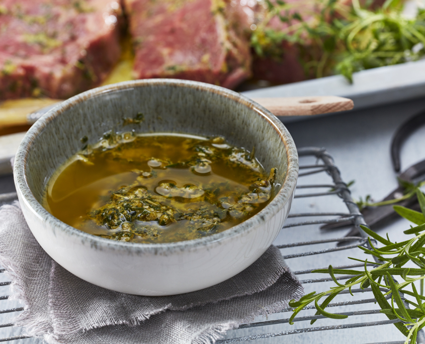 Eine kleine Schüssel mit einer Marinade aus grünen Kräutern steht auf einem Tuch auf einem Metallgitter zum Abkühlen. Im Hintergrund liegt ein mariniertes rohes Steak auf einer weißen Platte, perfekt eingefangen durch professionelle Food-Fotografie. Frische Kräuter sind überall verstreut und unterstreichen das rustikale kulinarische Ambiente.