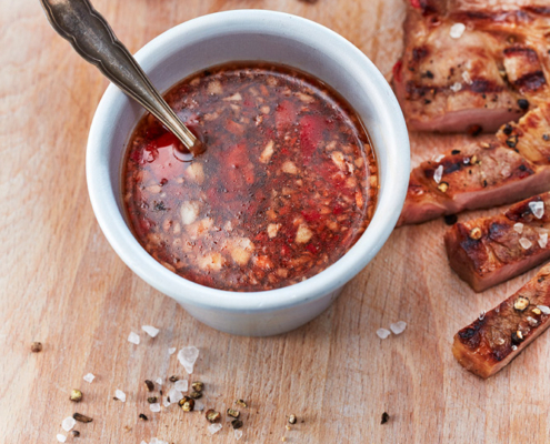 Eine kleiner weißer Porzellanbecher, gefüllt mit einer stückigen, rotbraunen Marinade, steht auf einem hölzernen Schneidebrett. Die Sauce weist sichtbare Gewürzreste und Chilliflocken auf. Neben dem Becher liegen ein Vintage-Löffel und gegrillte Fleischscheiben, die mit Salz und Pfeffer bestreut sind – eine perfekte Aufnahme für Food-Fotografie-Enthusiasten.
