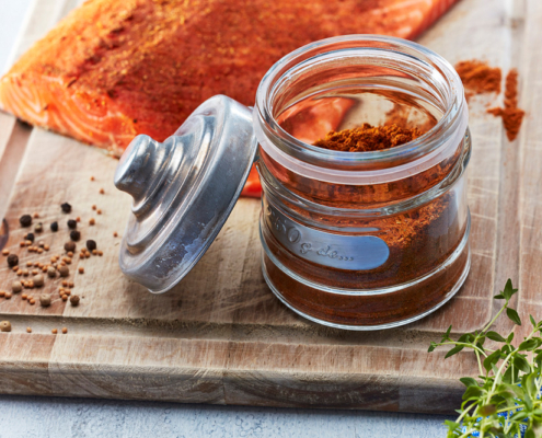 Ein hölzernes Schneidebrett mit einem Stück gewürztem Lachs steht neben einem Glas mit Cajun Gewürz ohne Deckel und verstreuten Gewürzsamen. Frische Kräuter sind kunstvoll in der Ecke platziert und zeigen exquisite Fähigkeiten eines Food-Fotografen für atemberaubende Fotografie von Lebensmitteln.