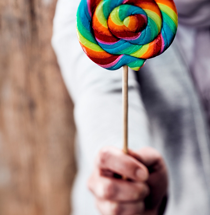 Eine Person in einem weißen Oberteil hält einen großen, bunten Lutscher in Spiralform. Der Hintergrund ist unscharf und hebt die leuchtenden, regenbogenfarbenen Süßigkeiten hervor – ein perfektes Beispiel exquisiter Foodfotografie.