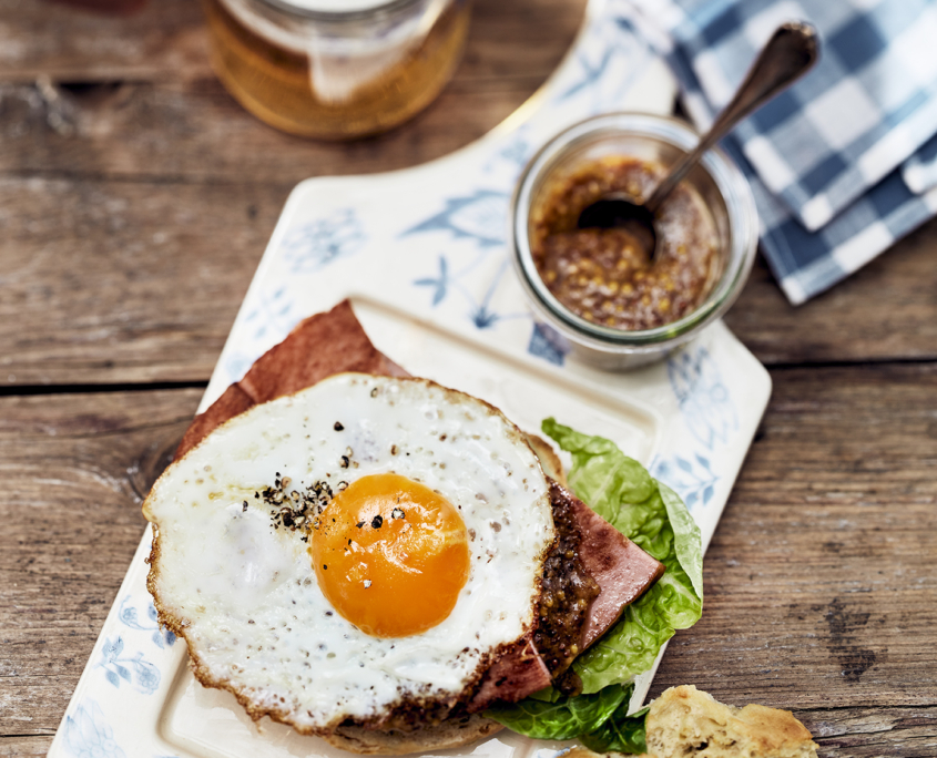 Ein köstliches belegtes Brötchen mit Schinken, einem perfekt gegarten Spiegelei obendrauf, Salat und Paprika, serviert auf einem weißen Teller mit einem rustikalen Porzellanbrett an der Seite. Aufgenommen von erfahrenen Food-Fotografen, umfasst diese Szene ein Glas körnigen Senf, schaumiges Bier in einem Krug und eine blaukarierte Serviette auf dem Holztisch.