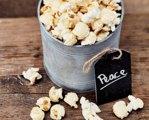 Ein mit Popcorn gefüllter Blecheimer steht auf einer rustikalen Holzoberfläche und verkörpert die Essenz der Foodfotografie. Auf einem schwarzen Schild am Eimer steht mit weißer Kreide „Peace“. Mehrere Stücke Popcorn sind verstreut und ergänzen die von einem erfahrenen Foodstylisten sorgfältig gestaltete Szene mit Holzbrettern als Hintergrund.