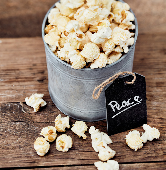 Ein mit Popcorn gefüllter Blecheimer steht auf einer rustikalen Holzoberfläche und verkörpert die Essenz der Foodfotografie. Auf einem schwarzen Schild am Eimer steht mit weißer Kreide „Peace“. Mehrere Stücke Popcorn sind verstreut und ergänzen die von einem erfahrenen Foodstylisten sorgfältig gestaltete Szene mit Holzbrettern als Hintergrund.