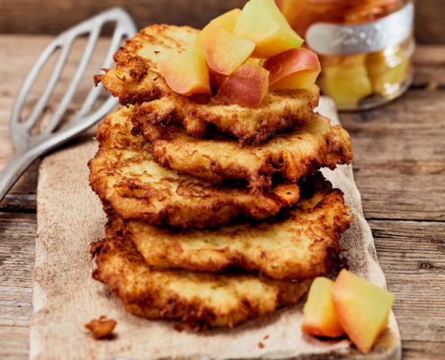 Ein Stapel goldbrauner Kartoffelpuffer, belegt mit Apfelwürfeln, liegt auf einem Holzschneidebrett und zeugt von tadellosen Fähigkeiten eines Foodstylisten. Ein Metallspatel steht daneben und ein Glasgefäß mit weiteren Apfelwürfeln vervollständigt die Bildsprache auf dem rustikalen Holztisch.