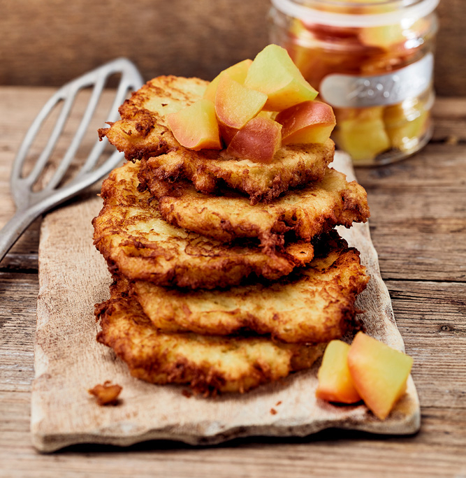 Ein Stapel goldbrauner Kartoffelpuffer, belegt mit Apfelwürfeln, liegt auf einem Holzschneidebrett und zeugt von tadellosen Fähigkeiten eines Foodstylisten. Ein Metallspatel steht daneben und ein Glasgefäß mit weiteren Apfelwürfeln vervollständigt die Bildsprache auf dem rustikalen Holztisch.