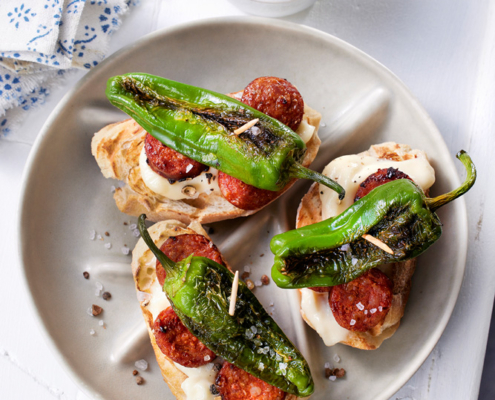 Auf einem weißen Teller ist ein Brotteller mit gegrillten grünen Paprikaschoten, Chorizo-Scheiben und cremiger Aioli angerichtet. In professioneller Foodfotografie-Manier aufgenommen, steht daneben eine kleine Schüssel mit noch mehr Aioli und einem Löffel. Der Tisch ist weiß und neben dem Teller liegt eine gemusterte Serviette.
