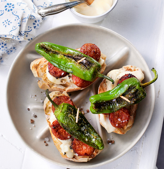 Auf einem weißen Teller ist ein Brotteller mit gegrillten grünen Paprikaschoten, Chorizo-Scheiben und cremiger Aioli angerichtet. In professioneller Foodfotografie-Manier aufgenommen, steht daneben eine kleine Schüssel mit noch mehr Aioli und einem Löffel. Der Tisch ist weiß und neben dem Teller liegt eine gemusterte Serviette.
