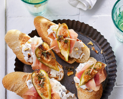 Ein runder schwarzer Teller mit vier Baguettescheiben, jede davon dekadent mit Feigen, Prosciutto und Streichkäse belegt, ist kunstvoll auf einer weißen Fläche arrangiert. Zwei grün getönte Gläser mit einem leichten Getränk und einem weißen Tuch ergänzen die raffinierte Bildsprache. Kunstvoll verstreute Krümel runden dieses Meisterwerk des Foodstylisten ab.