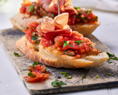 Zwei Scheiben geröstetes Brot, belegt mit geriebenen Tomaten und Kräutern, werden mit dünnen Scheiben Schinken garniert und mit Zahnstochern festgesteckt. Die von einem Foodstylisten perfekt gestylte Bruschetta wird auf einem rustikalen Holzbrett mit verstreuten Kräutern serviert.