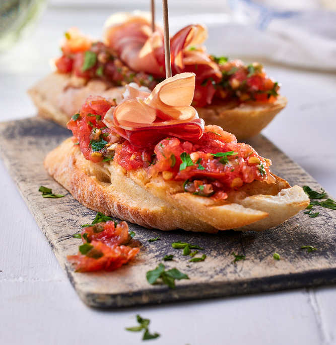 Zwei Scheiben geröstetes Brot, belegt mit geriebenen Tomaten und Kräutern, werden mit dünnen Scheiben Schinken garniert und mit Zahnstochern festgesteckt. Die von einem Foodstylisten perfekt gestylte Bruschetta wird auf einem rustikalen Holzbrett mit verstreuten Kräutern serviert.