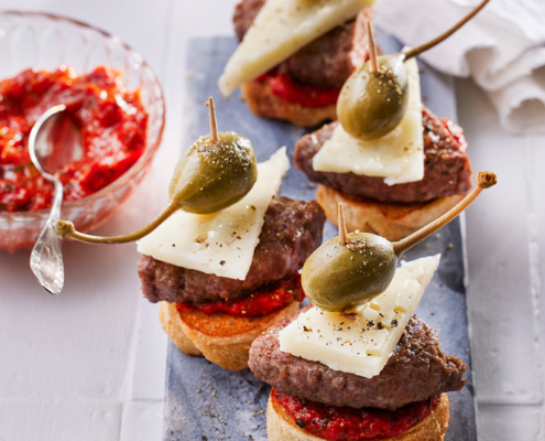 Auf einer weißen Fläche ist eine Schiefertafel mit vier Gourmet-Häppchen angeordnet. Jedes Häppchen besteht aus einer Scheibe Brot mit rotem Aufstrich, einem Stück Fleisch, einer Scheibe Käse, einer Kapernbeere und Gewürzen. Im Hintergrund sind eine Schale mit rotem Aufstrich und ein Löffel zu sehen, die exquisite Foodfotografie einfangen.