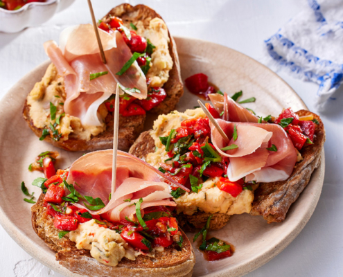 Drei Scheiben geröstetes Brot mit cremigem Aufstrich, Paprikasalsa, Kräutern und auf Zahnstochern aufgespießten Schinkenstücken, wunderschön angerichtet vom Foodstylisten auf einem weißen Teller. Im Hintergrund ist eine kleine Schüssel mit Paprikasalsa und Kräutern zu sehen, perfekt für begeisterte Foodfotografen.