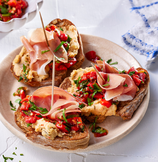 Drei Scheiben geröstetes Brot mit cremigem Aufstrich, Paprikasalsa, Kräutern und auf Zahnstochern aufgespießten Schinkenstücken, wunderschön angerichtet vom Foodstylisten auf einem weißen Teller. Im Hintergrund ist eine kleine Schüssel mit Paprikasalsa und Kräutern zu sehen, perfekt für begeisterte Foodfotografen.