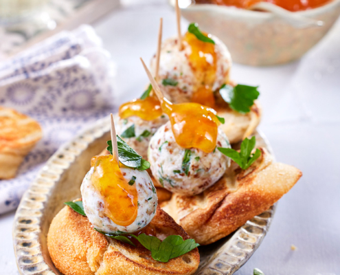 Ein Gericht mit gerösteten Baguettescheiben, belegt mit aufgespießten Ziegenfrischkäsebällchen, garniert mit einem Klecks Orangensauce und frischer Petersilie. Im Hintergrund sind eine kleine Schüssel Orangensauce und ein Glas Weißwein zu sehen, aufgenommen von erfahrenen Food-Fotografen. Eine gefaltete weiß-blaue Serviette rundet das Ganze ab.