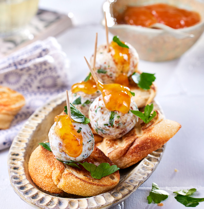 Ein Gericht mit gerösteten Baguettescheiben, belegt mit aufgespießten Ziegenfrischkäsebällchen, garniert mit einem Klecks Orangensauce und frischer Petersilie. Im Hintergrund sind eine kleine Schüssel Orangensauce und ein Glas Weißwein zu sehen, aufgenommen von erfahrenen Food-Fotografen. Eine gefaltete weiß-blaue Serviette rundet das Ganze ab.