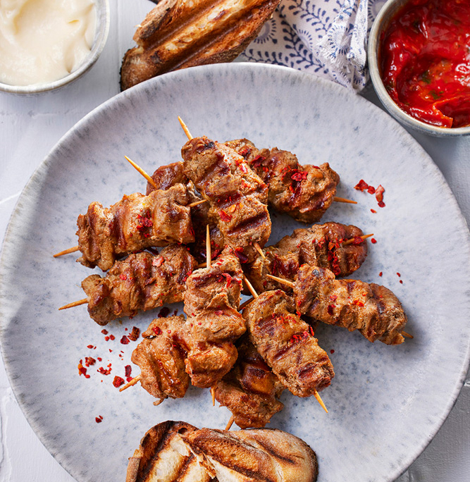 Ein Teller mit gegrilltem Fleischspieß, garniert mit roten Gewürzen, wird von gegrillten Baguettescheiben begleitet. Eine Schüssel Mayonnaise und eine Schüssel mit roter Soße stehen neben dem Teller auf einer weißen strukturierten Oberfläche, daneben ein gemustertes Tuch – eine perfekte Szene für Food-Fotografie, die exquisite Bildsprache einfängt.