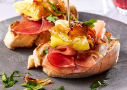 Zwei Scheiben Bruschetta mit Serano Schinken, gebratenem Ei, karamellisierten Zwiebeln und frischen Kräutern werden auf einem grauen Teller serviert. Ein Zahnstocher hält jeden Belag an Ort und Stelle. Die elegante Bildsprache fängt ein Glas Rotwein im Hintergrund ein und unterstreicht die kulinarische Kunstfertigkeit.