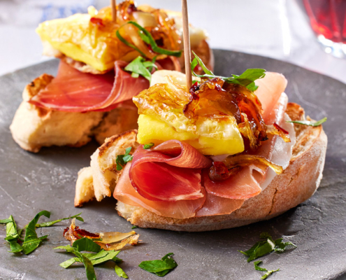 Zwei Scheiben Bruschetta mit Serano Schinken, gebratenem Ei, karamellisierten Zwiebeln und frischen Kräutern werden auf einem grauen Teller serviert. Ein Zahnstocher hält jeden Belag an Ort und Stelle. Die elegante Bildsprache fängt ein Glas Rotwein im Hintergrund ein und unterstreicht die kulinarische Kunstfertigkeit.