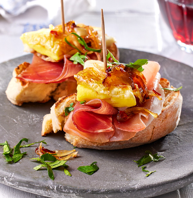 Zwei Scheiben Bruschetta mit Serano Schinken, gebratenem Ei, karamellisierten Zwiebeln und frischen Kräutern werden auf einem grauen Teller serviert. Ein Zahnstocher hält jeden Belag an Ort und Stelle. Die elegante Bildsprache fängt ein Glas Rotwein im Hintergrund ein und unterstreicht die kulinarische Kunstfertigkeit.