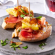 Zwei Scheiben Bruschetta mit Serano Schinken, gebratenem Ei, karamellisierten Zwiebeln und frischen Kräutern werden auf einem grauen Teller serviert. Ein Zahnstocher hält jeden Belag an Ort und Stelle. Die elegante Bildsprache fängt ein Glas Rotwein im Hintergrund ein und unterstreicht die kulinarische Kunstfertigkeit.