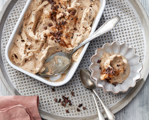 Eine rechteckige Schale mit cremigem Eis steht auf einem Metalltablett mit zwei silbernen Löffeln. Eine kleine, gewellte Schale auf dem Tablett ist perfekt für *Foodfotografie* und enthält eine Kugel Eis, garniert mit Schokoladenstückchen. Eine rosa Serviette liegt neben dem Tablett auf einer weißen, strukturierten Holzoberfläche.