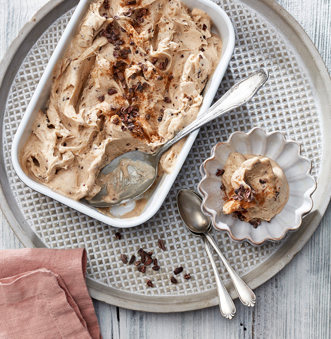 Eine rechteckige Schale mit cremigem Eis steht auf einem Metalltablett mit zwei silbernen Löffeln. Eine kleine, gewellte Schale auf dem Tablett ist perfekt für *Foodfotografie* und enthält eine Kugel Eis, garniert mit Schokoladenstückchen. Eine rosa Serviette liegt neben dem Tablett auf einer weißen, strukturierten Holzoberfläche.