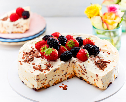 Eine dekadente Eistorte, gefertigt von einer talentierten Foodstylistin, ist mit frischen Erdbeeren und Brombeeren belegt und steht auf einem weißen Tortenständer. Ein Stück wurde entfernt, so dass das attraktive Innere mit Schokoladenraspeln zum Vorschein kommt. Im Hintergrund sind eine Vase mit bunten Blumen und ein Teller mit einem Stück Torte zu sehen – ein wahres Meisterwerk der Foodfotografie.